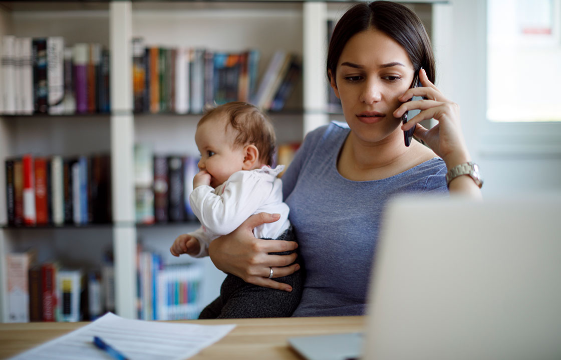 woman holding a baby while on her cell phone and sitting in front of a computer